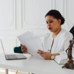 Femme en blouse blanche lit des documents à un bureau avec ordinateur et statue en bronze