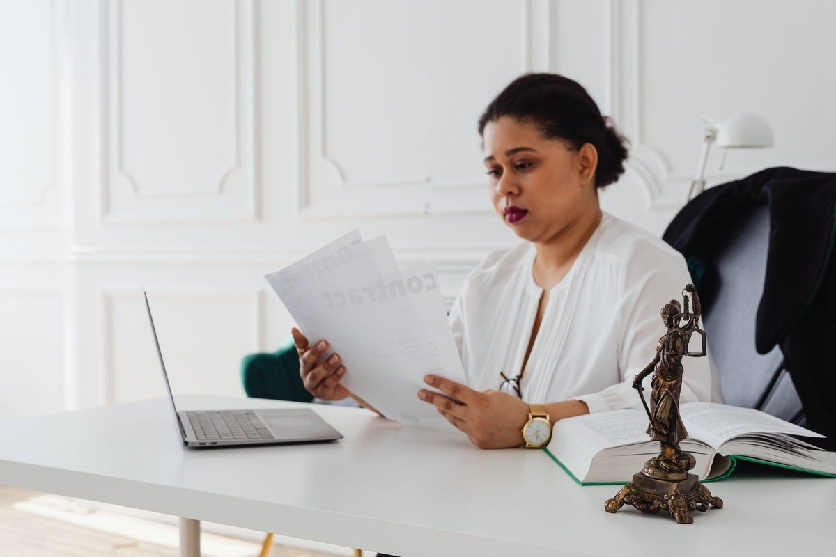 Femme en blouse blanche lit des documents à un bureau avec ordinateur et statue en bronze