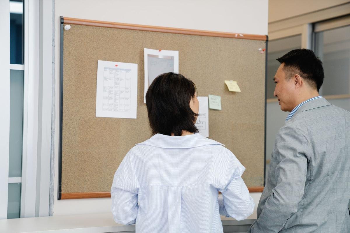 Deux personnes regardent un panneau d'affichage en liège avec documents épinglés sur un mur blanc de bureau Deux personnes regardent un panneau d'affichage en liège avec documents épinglés sur un mur blanc de bureau