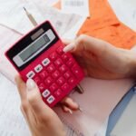Des mains manipulent une calculatrice rouge au-dessus de documents et d'un crayon sur une table